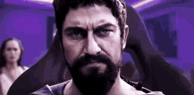 a man with a beard is sitting in a chair and looking at the camera with a woman in the background .