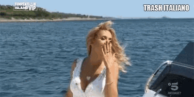 a woman in a white dress is standing on a boat in the ocean covering her mouth .