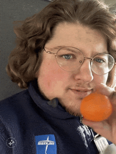 a man wearing glasses holds an orange in his hand and has a patch on his jacket that says ' hiking '