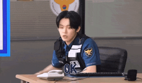 a young man in a police uniform is sitting at a desk with a phone and a keyboard .