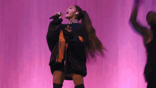 ariana grande is singing into a microphone on stage in front of a pink background .