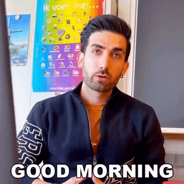 a man with a beard is sitting in front of a chalkboard and saying good morning .