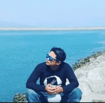 a young man wearing sunglasses is sitting on a rock near the water .