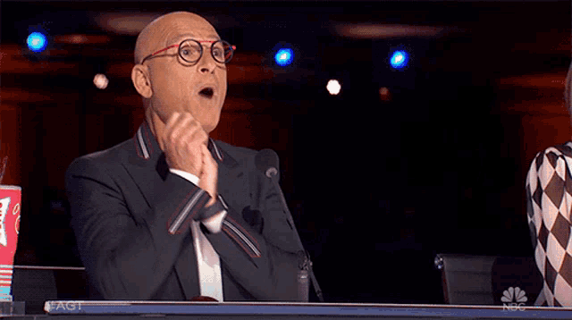 a man in a suit and glasses sitting in front of a microphone with his mouth open and a coca cola cup in front of him