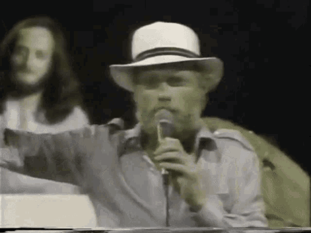 a man with a beard is singing into a microphone while wearing a hat
