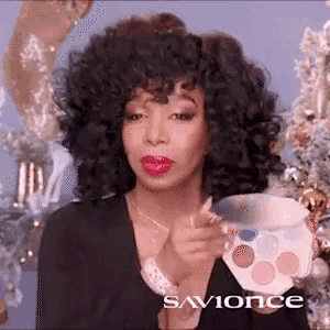 a woman with curly hair is holding a makeup palette in her hand .