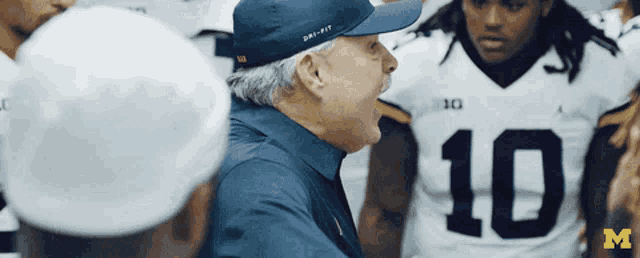 a man wearing a dri-fit hat stands in front of a group of football players