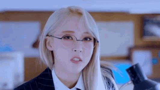 a woman with blonde hair wearing glasses and a suit looks at the camera