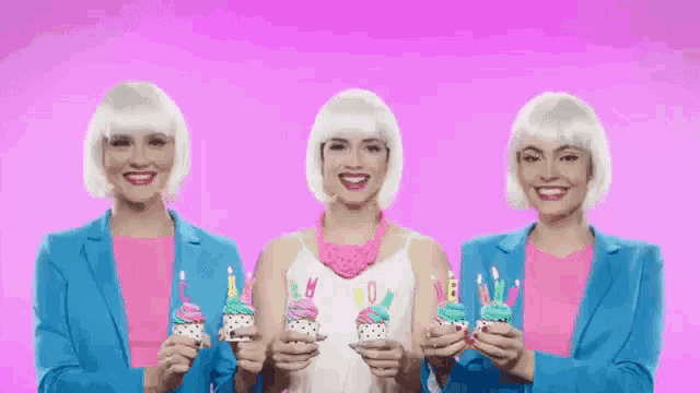 three women in white wigs are holding cupcakes with birthday candles .