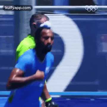 a man in a blue shirt is running on a track with a man in a green shirt behind him .