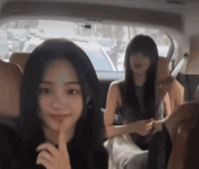 two women are sitting in the back seat of a car . one of the women is holding her finger to her lips .