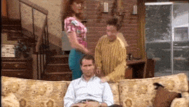 a man is sitting on a couch in a living room while a woman stands next to him .