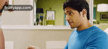 a man in a blue shirt is sitting at a table in a kitchen .
