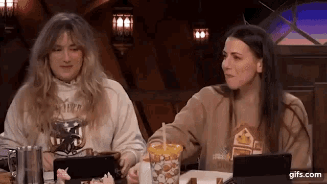 two women are sitting at a table with a cup of popcorn .