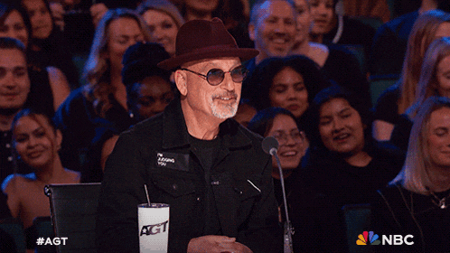 a man wearing a hat and sunglasses applauds in front of a crowd with a nbc logo behind him