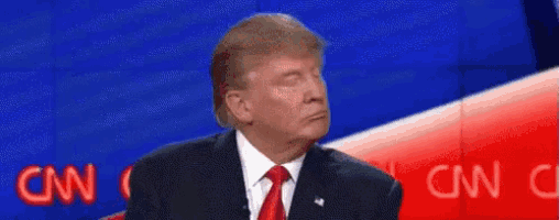 donald trump is wearing a suit and tie and making a funny face while sitting on a stage .