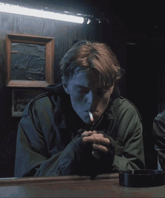 a man in a green jacket smoking a cigarette next to an ashtray
