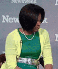a woman in a green dress and yellow cardigan is sitting in front of a newsweek sign