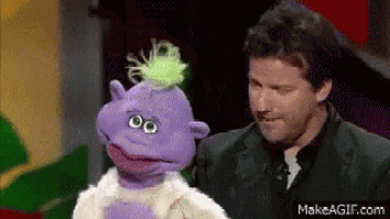 a man in a suit is standing next to a purple puppet with green hair