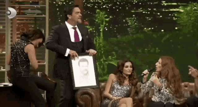 a man in a suit and tie holds a framed picture of a man while two women sing into microphones