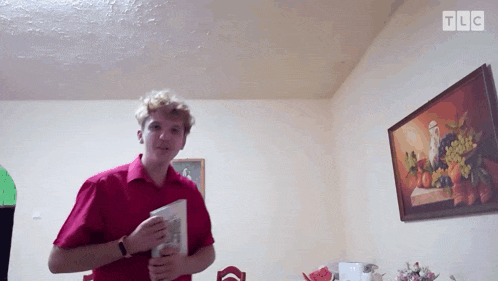 a man in a red shirt is holding a book in front of a framed painting with tlc written on it