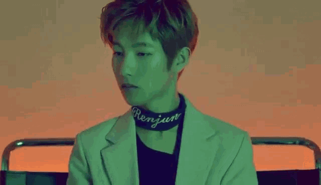 a young man is sitting in a chair wearing a green jacket and a choker .