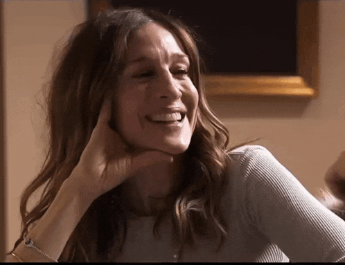 a woman wearing a grey sweater is smiling with her hand on her chin