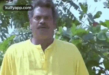 a man with a mustache is wearing a yellow shirt and standing in front of a tree .