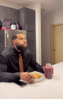 a man in a black shirt and tie is sitting at a table eating a bowl of food and drinking a smoothie .