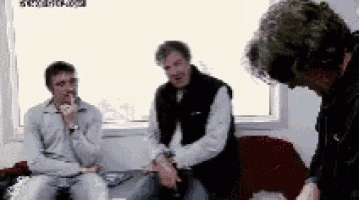 three men are sitting on a couch in front of a window and one of them is smoking a cigarette .