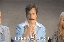a man with a mustache is sitting in front of a group of people and says `` keep smiling ! ''