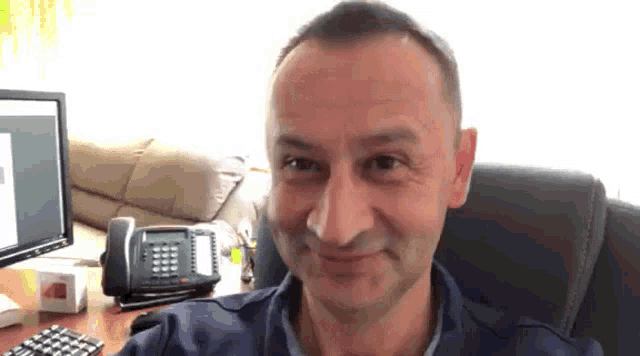 a man is smiling while sitting at a desk in front of a computer and a phone .