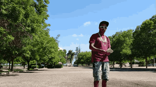 a man wearing sunglasses and a maroon shirt holds a bunch of money in front of trees