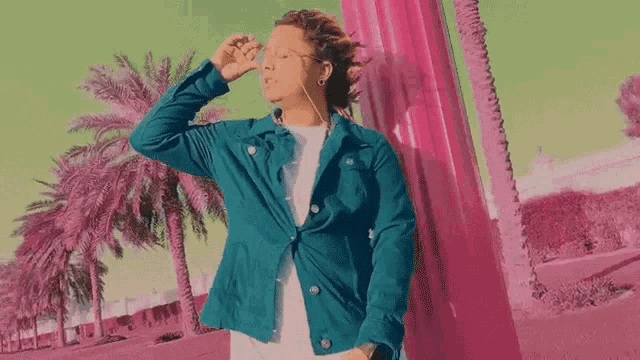 a man wearing a blue jacket and headphones is standing in front of a pink pillar and palm trees .