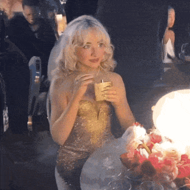 a woman in a gold dress is sitting at a table holding a drink