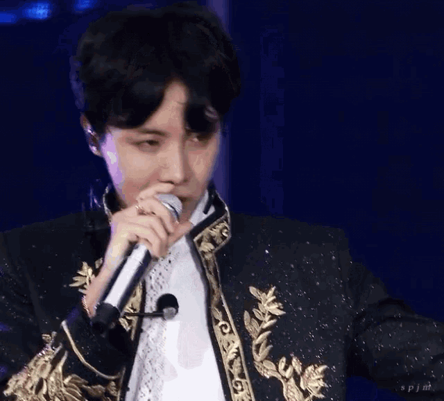 a man is singing into a microphone while wearing a black jacket with gold trim