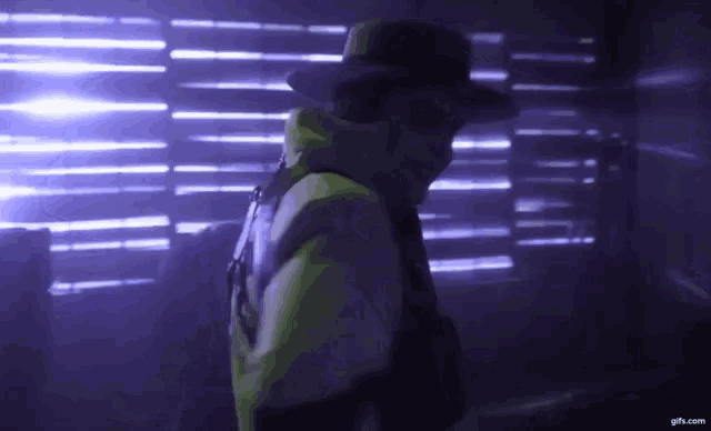 a man wearing a hat is standing in a dark room with purple lights behind him
