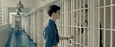 a man in a blue shirt and tie is standing in a jail cell talking to a prisoner behind bars .