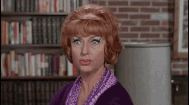 a woman with red hair is wearing a purple sweater and a purple scarf in front of a bookshelf .