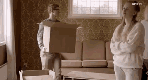 a man is carrying a cardboard box in a living room while a woman stands behind him .