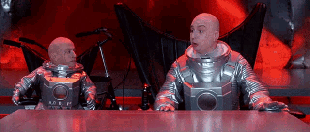 two bald men in space suits are sitting at a table with a box that says h2o2