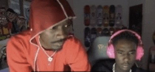a man in a red hoodie and headphones is sitting next to another man in a room .