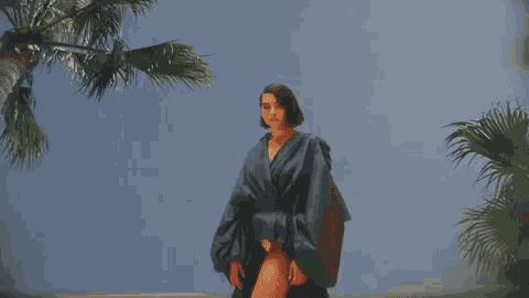 a woman in a blue kimono is standing in front of a palm tree on the beach .