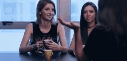 two women are sitting at a table with a cup of coffee and talking .