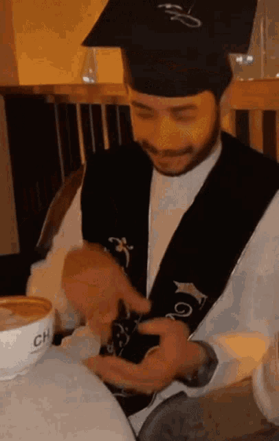 a man wearing a graduation cap and gown is sitting at a table with a cup of coffee