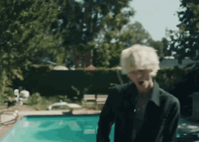a man with blonde hair is standing next to a swimming pool in a backyard .