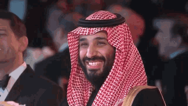 a man in a keffiyeh is smiling at the camera while sitting in a crowd of people .