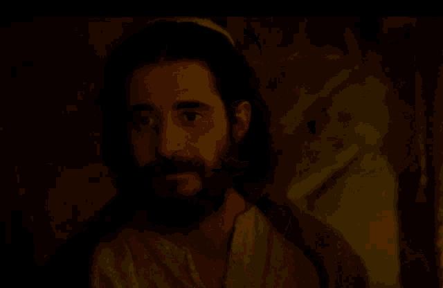 a man with long hair and a beard looks at the camera in a dark room