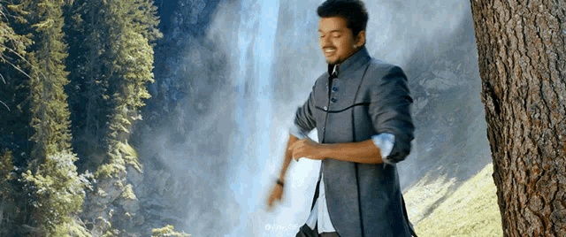 a man in a suit leans against a tree in front of a waterfall