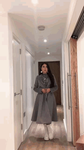 a woman wearing a grey coat and white boots is walking down a hallway